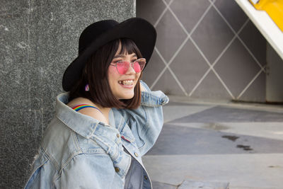 Side view of smiling young woman wearing sunglasses and hat standing against wall