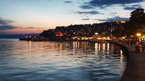 Illuminated city by sea against sky at sunset