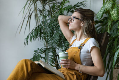 Side view of woman with book holding coffee while sitting on chair