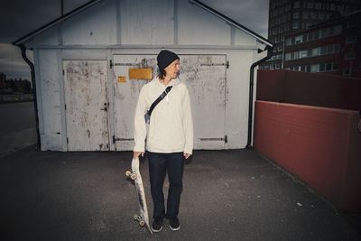 Man standing with skateboard against shed on street