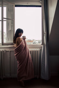 Side view of woman wrapped in blanket having coffee while standing by window