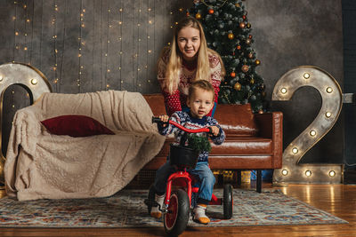 Portrait of son riding tricycle with mother against christmas tree