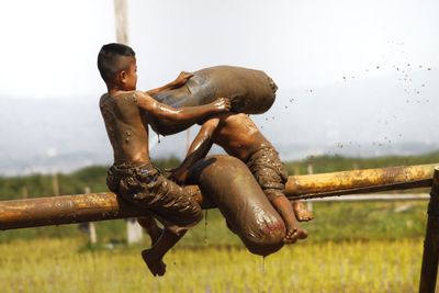 Messy shirtless boys fighting while sitting on bamboo against sky
