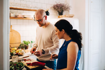 Woman with wineglass looking at male friend cutting pepper in kitchen