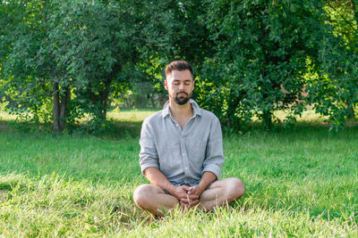 Portrait of young handsome european man in casual clothing sitting on a grass in summer park.