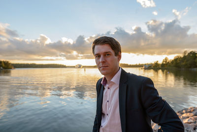 Portrait of young man standing by lake against sky during sunset