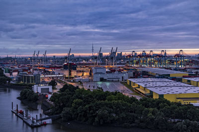 Cranes at harbor against cloudy sky in city during sunset