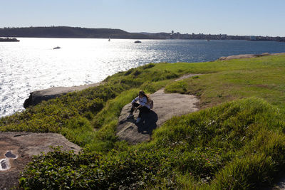 Rear view of man sitting on shore by sea against sky