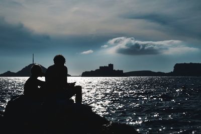 Rear view of man sitting on rock by sea against sky during sunset