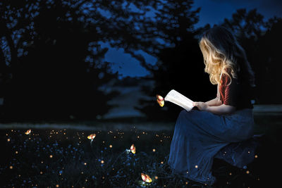 Woman reading book while sitting on grass against trees at night