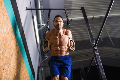 Shirtless male athlete exercising on gymnastic rings in gym