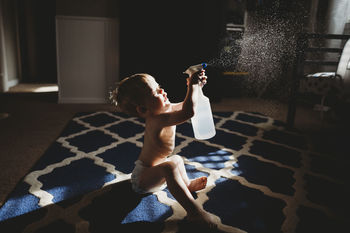 Side view of shirtless baby boy spraying water while sitting on carpet at home