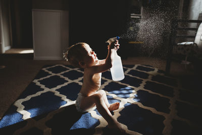 Side view of shirtless baby boy spraying water while sitting on carpet at home