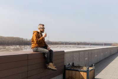 Full length of man drinking smoothie sitting on wall against sky