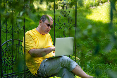 Middle aged man with laptop working outside in garden, green home office concept
