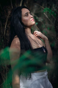 Beautiful woman looking away standing by tree trunk in forest