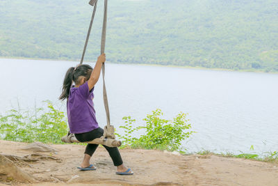 An asian girl is sitting on a swing tied to a tree, behind a mountain river.