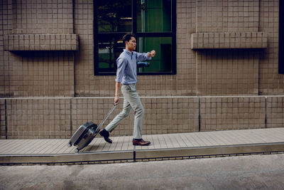 Side view of young man with suitcase checking time while walking on sidewalk