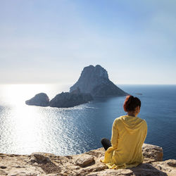 Young woman in yellow zip jacket sitting on a cliff looking to famous es vedra