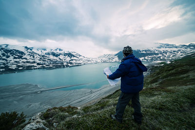 Side view of mature man holding map while standing at lakeshore against cloudy sky during winter