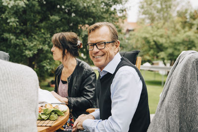 Portrait of smiling senior man sitting with friends at dining table in back yard
