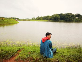 Man sitting on grass by lake against sky
