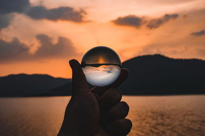 Human hand holding crystal ball against sky during sunset