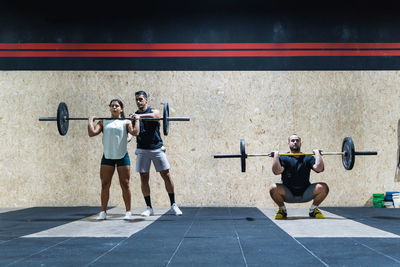 Full body group of strong hispanic athletes lifting heavy barbells near wall in light gym during weightlifting training together