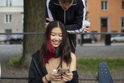 Teenage boy looking at smiling female friend using smart phone in park