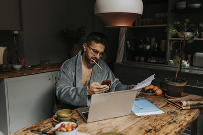 Man holding bills using smart phone while sitting with laptop on dining table at home