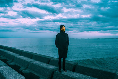 Full length of person wearing mask while standing on retaining wall by sea against cloudy sky during sunset