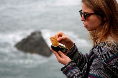 Side view of young woman holding sunglasses at beach