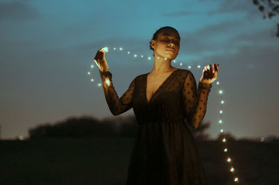 Beautiful woman with holding string lights outdoors at dusk