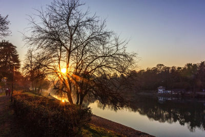 Bare trees by lake against sky during sunset