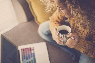 Woman holding coffee cup looking into laptop in living room at home