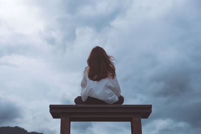 Rear view of woman sitting on bench against cloudy sky