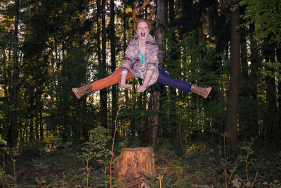 Full length portrait of happy woman jumping over tree stump in forest