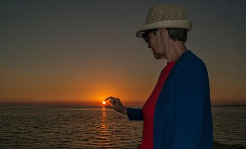 Optical illusion of woman holding sun while standing by sea during sunset