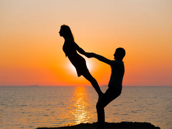 Silhouette couple exercising at beach against sky during sunset