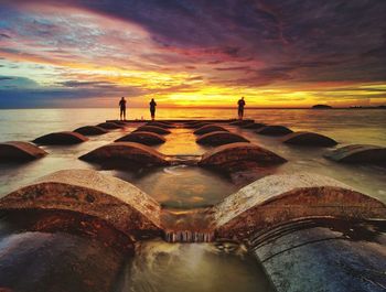 People standing on pipes at beach against sky during sunset