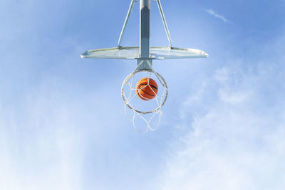 From below of orange basketball falling into round hoop with net placed against blue sky on sunny summer day in city