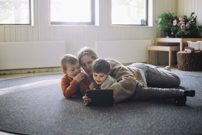 Female teacher watching digital tablet with boys while lying down on carpet at preschool