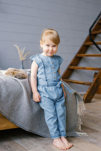 A cute two-year-old girl in a denim jumpsuit with bare feet stands by the bed at home