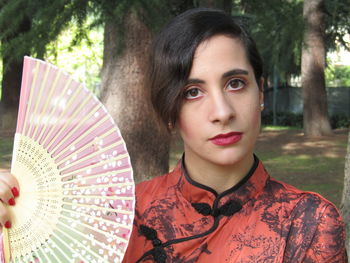 Close-up portrait of woman holding hand fan while standing against trees