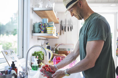 Side view of man washing cherry tomatoes under faucet in kitchen at home