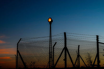 Illuminated lighting equipment by silhouette fence against sky