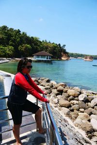 Side view of young woman in sunglasses standing by railing at rocky beach