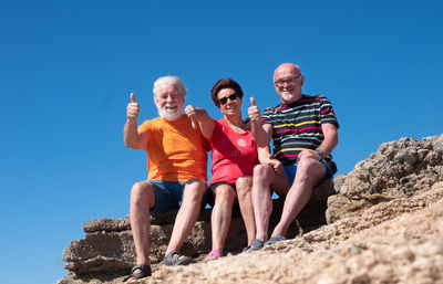 Low angle view portrait of senior friends gesturing while sitting on rock against sky