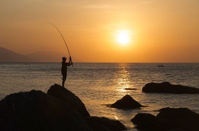 Silhouette person standing on rock by sea against sky during sunset