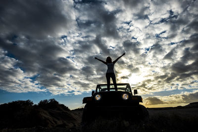 Low angle view of woman with arms raised standing on off-road vehicle at sunset